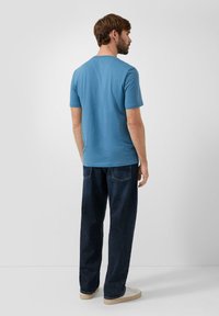 Niebieska bawełniana koszulka z krótkim rękawem i okrągłym dekoltem, zestawiona z ciemnymi dżinsami i białymi trampkami, pokazana od tyłu.