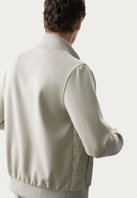 Giacca bomber grigio chiaro con una texture morbida e liscia, polsini a coste e un'accattivante zip laterale. Presenta un design semplice e minimalista.