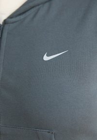 Top deportivo de Nike en tela gris oscuro, con una textura suave y un logo blanco en el pecho, cierre con cremallera y diseño aerodinámico.