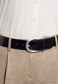 Ceinture en cuir noir avec boucle ovale en argent portée sur un pantalon beige texturé et une chemise crème à boutons rentrée.
