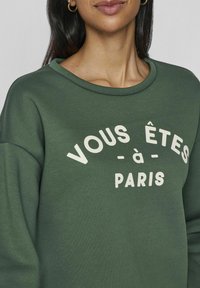 Sudadera verde con cuello redondo, con texto blanco que dice "VOUS ÊTES À PARIS." Textura suave y corte relajado.