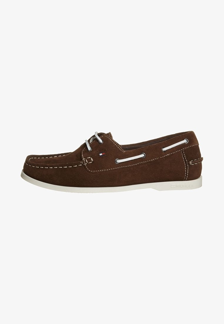 Braune Wildleder-Bootschuhe mit weißen Nähten und Schnürsenkeln, ausgestattet mit seitlichen Ösen und einer weißen Gummisohle. Logodetail in der Nähe der Ferse.