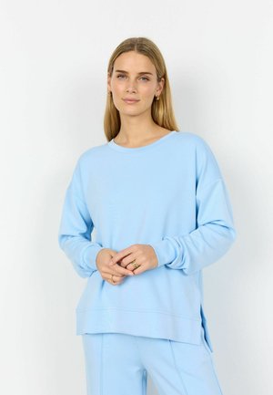 Junge Frau mit blondem Haar, die ein hellblaues Langarmshirt und passende Hose trägt, vor einem schlichten weißen Hintergrund stehend.