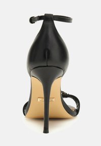 Schwarze high-heeled Sandale mit einem Knöchelriemen, dünnem Stiletto-Absatz und beigem Fußbett, von hinten auf einem weißen Hintergrund abgebildet.