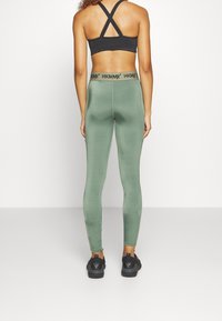 Zielone legginsy wykonane z gładkiej, elastycznej tkaniny z wysokim pasem z czarnym logo. Noszone z czarnym sportowym biustonoszem i czarnymi trampkami.