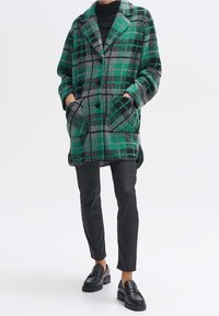 Person in einem langen grün-grauen karierten Mantel mit schwarzen Knöpfen, schwarzem Rollkragenpullover, schwarzen schmalen Hosen und schwarzen Slippern, die die Hände in den Taschen hat.