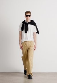 Camiseta branca, calças largas em tom claro, suéter preto pendurado nos ombros e óculos de sol pretos. Modelo a usar sapatos pretos, em pé contra um fundo claro.