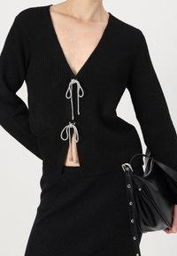 Černý pletený svetr s hlubokým výstřihem do V, s stříbrnými šňůrkami na stažení v pase a dlouhými rukávy. Strukturovaný střih.