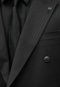 Μαύρο σακάκι με ρίγες, με μαύρο πουκάμισο και γραβάτα, που έχει ένα ασημένιο κυκλικό καρφίτσα στο αριστερό πέτο.