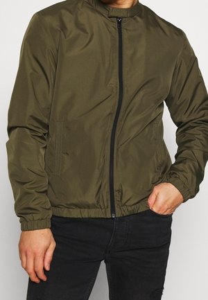 Homme portant une veste zippée vert olive avec des poignets élastiques et un pantalon noir, une main dans la poche, recadré au niveau du cou et des cuisses.