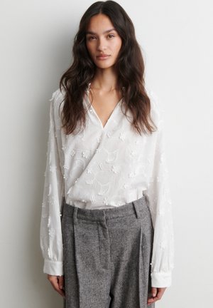 Femme aux longs cheveux foncés portant un chemisier blanc brodé de fleurs et un pantalon gris taille haute sur un fond uni.