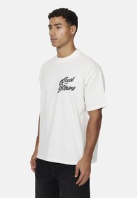 Biała bawełniana koszulka z krótkimi rękawami i okrągłym dekoltem. Z przodu znajduje się czarny napis "A Good For Nothing". Prosty, luźny krój.