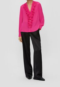 Modellen bär en ljusrosa blus med volanger och knappar, matchad med svarta vida byxor och spetsiga svarta skor, poserande mot en neutral bakgrund.