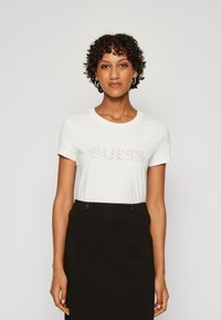 Camiseta blanca de manga corta con "GUESS LOS ANGELES" en strass. Combina con una falda negra de talle alto con detalles de botones.
