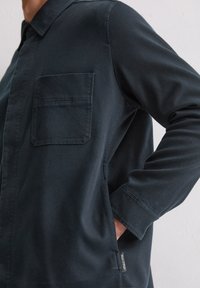 Dunkel marineblaues Hemd aus einem weichen Stoff, mit einer Brusttasche, langen Ärmeln und einem Seitenschlitz. Etikett am Saum sichtbar.