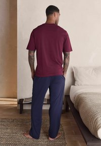 Bordowa koszula z krótkim rękawem w połączeniu z granatowymi spodniami. Koszula ma okrągły dekolt; spodnie są luźne. Boso na teksturowanym dywanie.