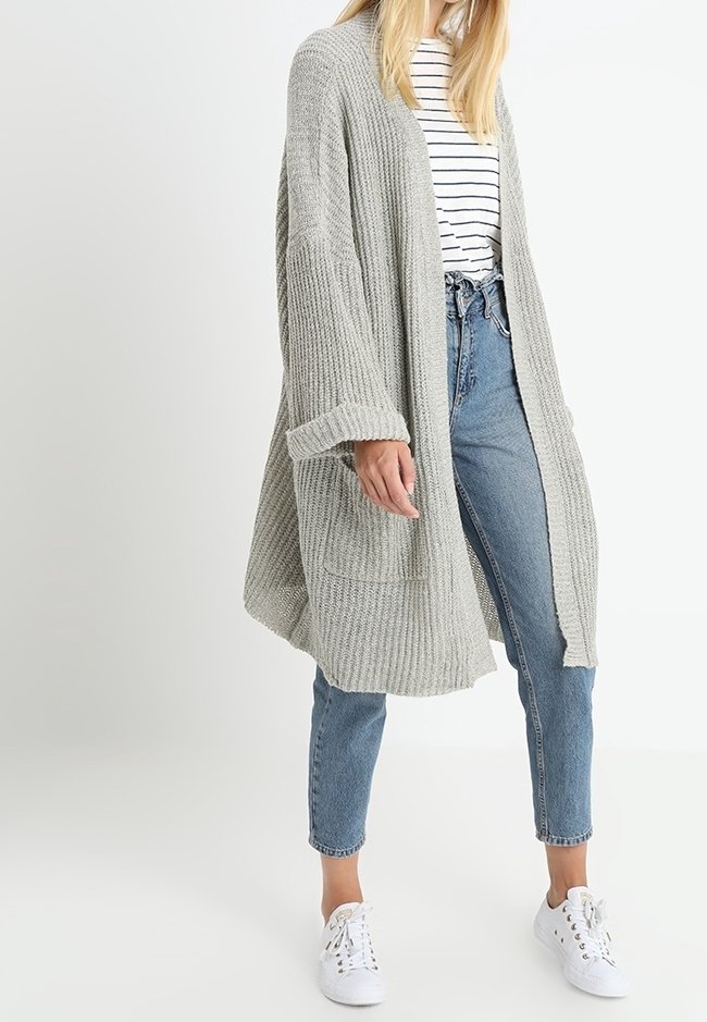 Cardigan ample gris clair à texture côtelée, devant ouvert et manches longues, porté sur une chemise rayée, associé à un jean bleu et des baskets blanches.