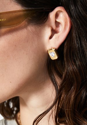 Boucles d'oreilles géométriques en or avec une pierre précieuse transparente en forme de cœur. Fini métallique lisse avec des lignes épurées, portées sur le lobe de l'oreille. Cheveux partiellement visibles.