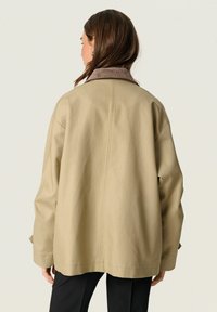 Manteau beige à coupe décontractée, avec col en velours côtelé et manches longues. Présente une couture arrière piquée et une texture de tissu douce.