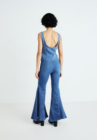 Denim kombinezon sa dubokim izrezom na leđima, proširenim nogavicama i prilagođenim korzetom. Materijal je lagani plavi pamuk s minimalnim detaljima šivanja.