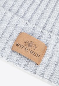 Cappello lavorato a maglia di colore grigio chiaro con trama a coste e targhetta in pelle marrone con la scritta "WITTCHEN" in rilievo e elementi decorativi.