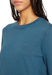 Türkisfarbenes Langarmshirt mit einem gerippten Ausschnitt und einem kleinen, gestickten Logo auf der Brust. Der Stoff wirkt weich und strukturiert.