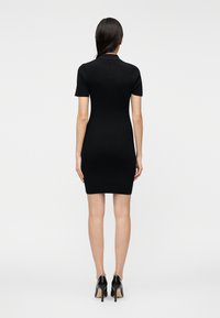 Črna ribana obleka s kratkimi rokavi, oprijetega kroja, z ovratnikom in dolžino nad koleni. Oblečena s črnimi visokimi petami.