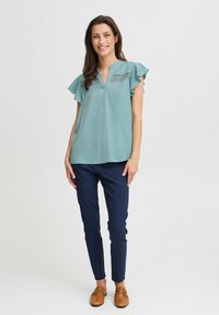 Blouse teal à manches courtes avec des manches volantes et un détail en dentelle au niveau du col, associée à un pantalon slim-fit bleu marine et des chaussures à enfiler marron.