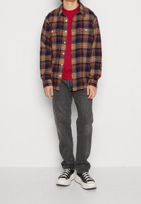 Chemise en flanelle à carreaux bleus, marron et jaunes, avec deux poches poitrine, superposée sur un T-shirt rouge. Assortie avec un jean gris et des baskets noires.
