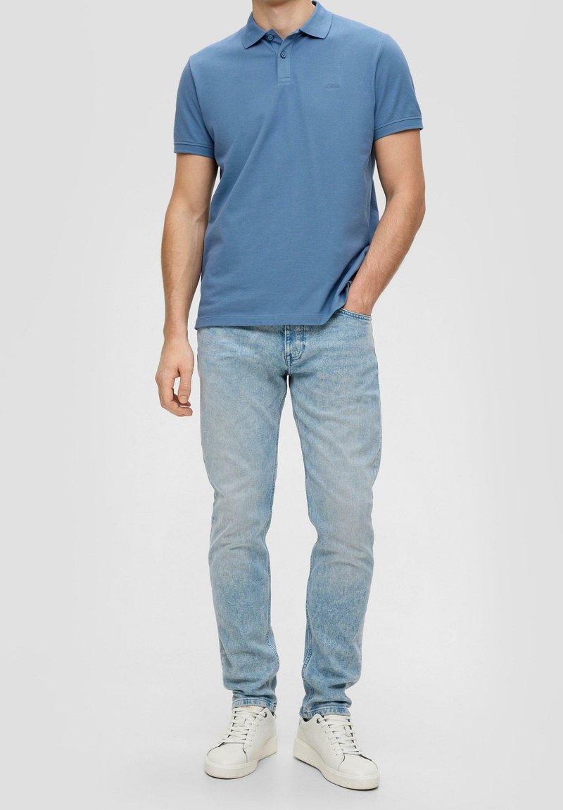 Homme portant un polo bleu à manches courtes, un jean bleu clair et des baskets blanches, debout avec une main dans la poche sur un fond uni.