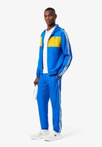 Blauwe trainingsbroek met gele accenten, ritsjack, bijpassende broek, wit logo op de zijkanten, gecombineerd met witte sneakers en een fles in de hand.