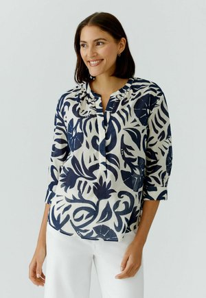 Femme souriante, portant un chemisier blanc avec un imprimé botanique bleu foncé audacieux et un pantalon blanc, debout devant un fond clair uni.