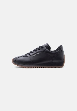 Black leather sneaker with brown ridged sole, lace-up closure, and "MICHAEL KORS" logo on side.