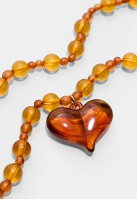 Bernsteinfarbene Perlenkette mit einem großen, durchscheinenden Herzanhänger mit wirbelnden Mustern. Glatte Textur, runde Perlen, minimaler Metallschmuck.