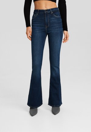 Femme portant un jean évasé bleu foncé, un haut court à manches longues noir et des chaussures grises pointues, debout devant un fond uni.