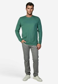 Grünes Langarmshirt mit Rundhalsausschnitt, kombiniert mit grauer Hose. Das Modell trägt weiße Sneaker, Hände in den Taschen, neutraler Hintergrund.