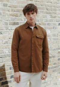 Hnedá košeľa na gombíky vyrobená z bavlny, s jedným vreckom na hrudi a klasickým golierom, v kombinácii s svetlými nohavicami.