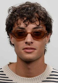 Gafas de sol tintadas en marrón con forma ovalada y montura transparente, llevadas sobre piel clara. Mostrando un suéter suave y texturizado con rayas horizontales.