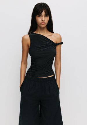 Top nero asimmetrico con dettaglio annodato sulla spalla, realizzato in tessuto morbido e elastico; indossato con culotte scure e morbide.