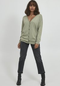 Cardigan oversize verde chiaro con scollo a V, chiusura a bottoni e maniche lunghe, abbinato a jeans scuri e stivaletti neri alla caviglia.