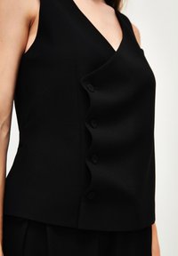 Top senza maniche nero con scollatura a V, caratterizzato da un pannello ondulato decorato con cinque bottoni neri, realizzato in un tessuto morbido e strutturato.