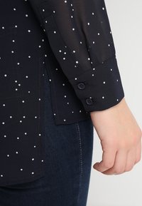 Mörkblå blus med vita prickar, transparent tyg, knäppt manschett och lätt struktur. Bärs med mörka jeans för en lagerpå-lager look.