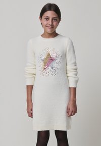Ragazza che indossa un vestito bianco a maniche lunghe con una grande stella fatta di paillette dorate, rosa e argentate sul davanti, in piedi contro uno sfondo semplice.