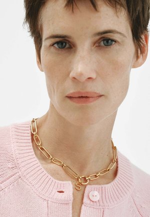 Mujer con cabello corto y castaño lleva un collar de cadena dorada con un colgante en forma de "S" y un suéter de punto rosa con un botón cerca del cuello.