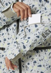 Jasnoszara kurtka z wielokolorowym, specklowanym wzorem, kieszenią na zamek na ramieniu oraz regulowanymi mankietami. Posiada gładki materiał i teksturowane akcenty.