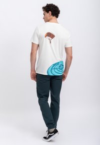 T-shirt bianca in cotone con una grafica di paracadutista sulla schiena e un design circolare blu all'orlo. Abbinata a pantaloni verde scuro e scarpe nere.