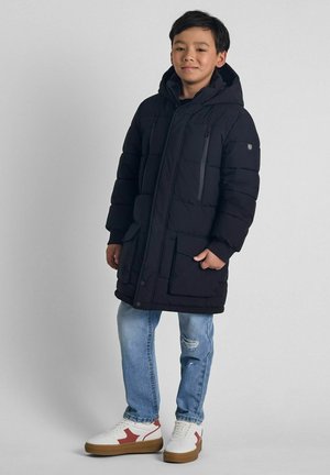 Černá péřová bunda s kapucí, s předním zipem, bočními kapsami a texturovaným povrchem. Nosená přes světle modré džíny a bílé sneakers.