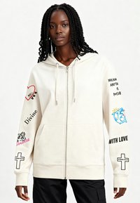 Jasnobeżowa bluza z kapturem zapinana na zamek, wykonana z miękkiego materiału. Na rękawach znajdują się nadruki graficzne, w tym teksty i ilustracje. W komplecie przednie kieszenie.