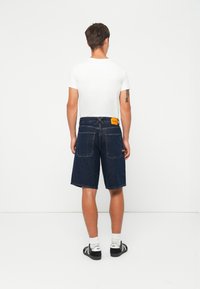 Pantalones cortos de mezclilla azul oscuro para hombre con un corte recto, dos bolsillos traseros y una etiqueta amarilla en la cintura. Se llevan con una camiseta blanca y zapatos negros.