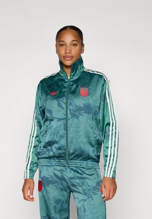 Giacca di tuta in satin verde con motivo floreale, maniche a righe bianche e accenti rossi del logo. Collo alto e chiusura frontale con zip.